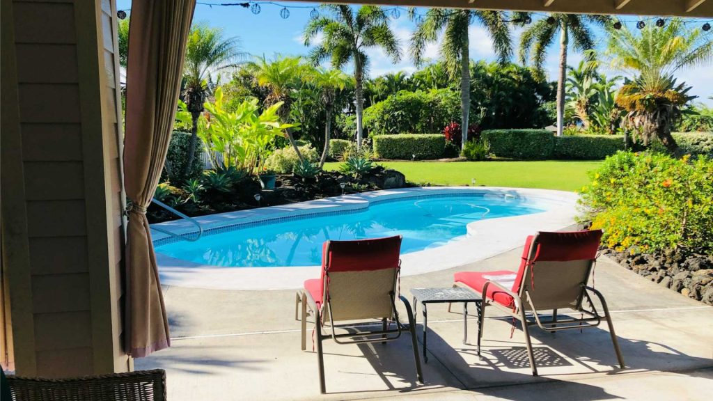 A clean swimming pool with turquoise blue water in a beautifully landscaped backyard with two red lounge chairs and a table. contact us